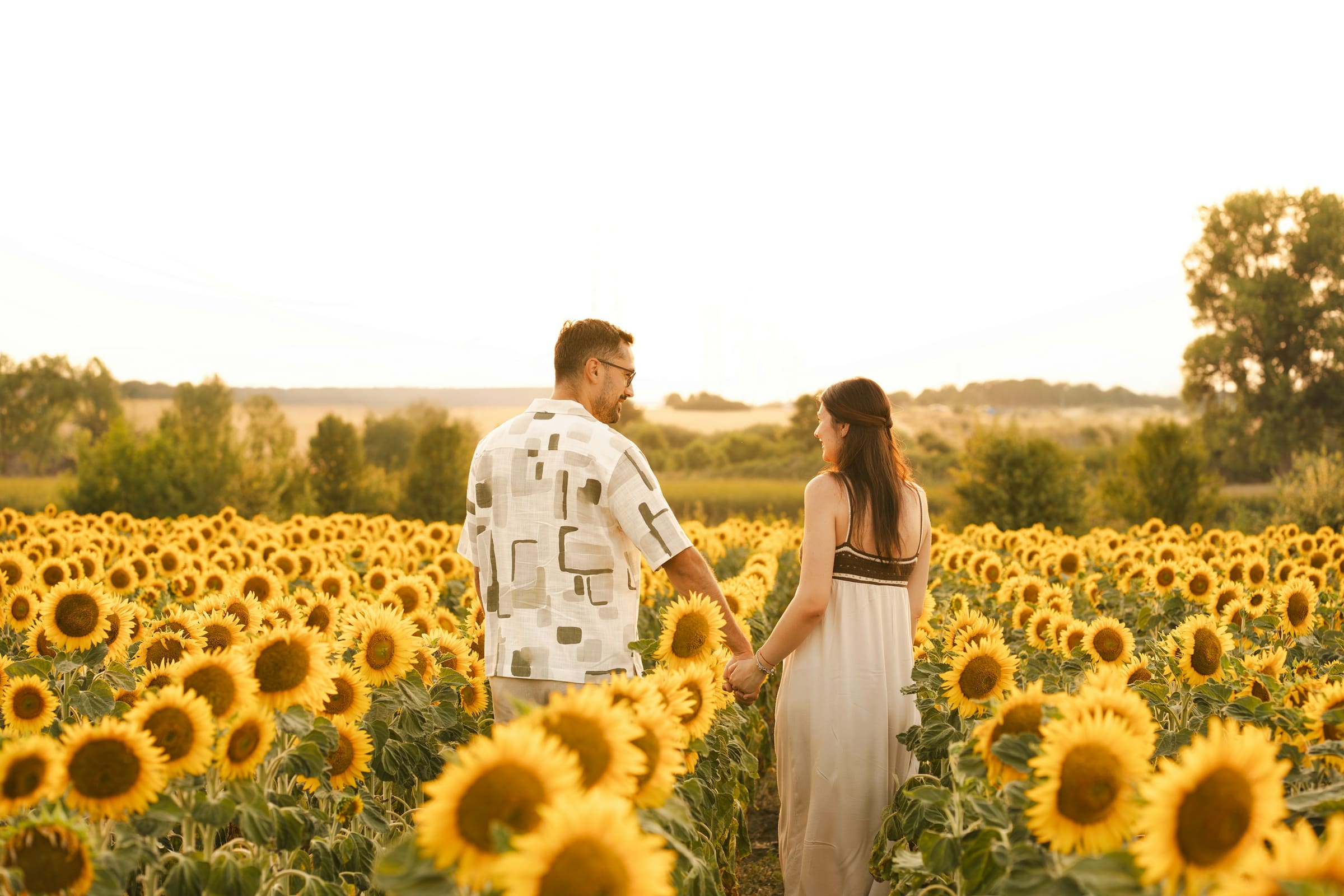 Couple posing in a sunflower field at golden hour, woman in white dress and man in patterned shirt facing each other